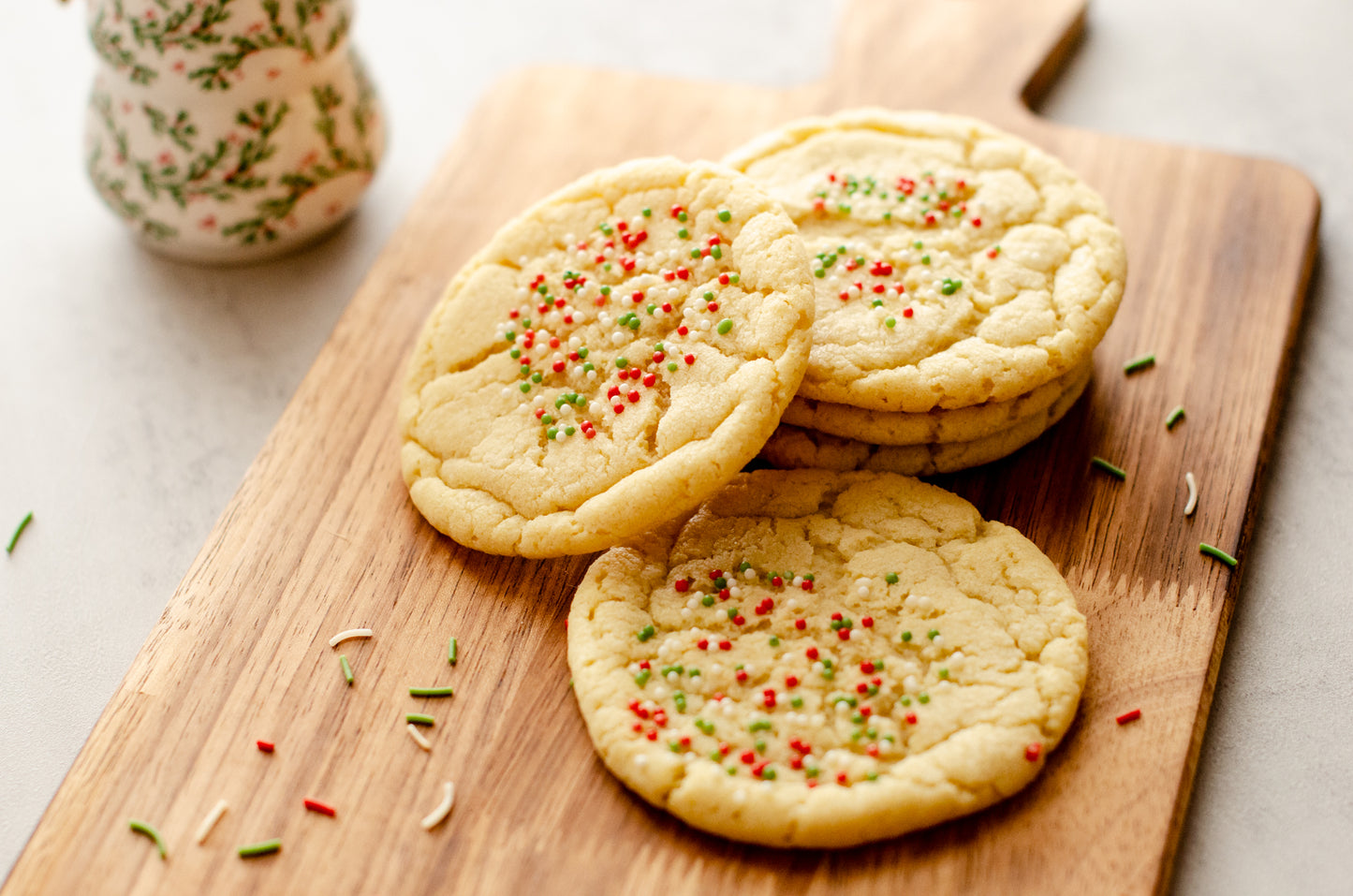 Three holiday themed sugar cookies with festive sprinkles on a wooden board with a decorative Christmas tree ornament in the background, baked fresh by Chillable Bake Shop in Darien, Wisconsin.