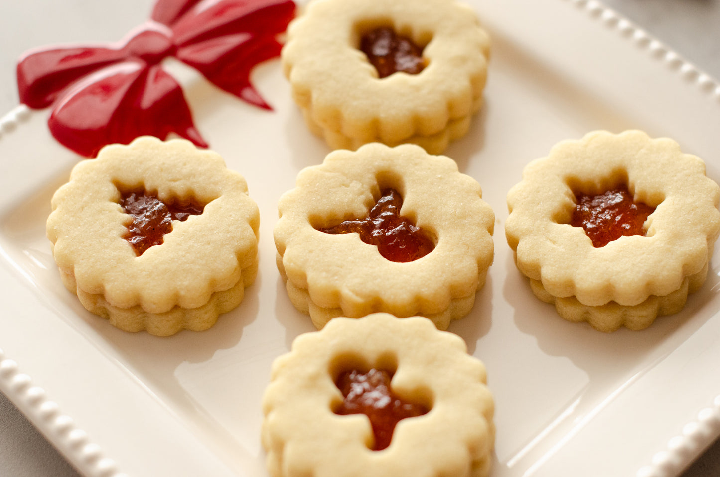 Peach nutmeg shortbread Linzer cookies with cutout centers on a white plate with a red bow, on a gray surface with Christmas decorations. Baked fresh by Chillable Bake Shop in Darien, WI.