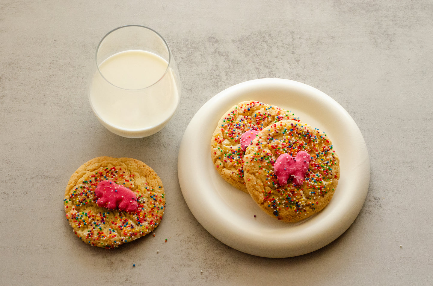 Three sugar cookies, each topped with a pink frosted animal cookie and colorful sprinkles on a white plate, accompanied by a glass of milk, baked fresh by Chillable Bake Shop in Darien, Wisconsin.