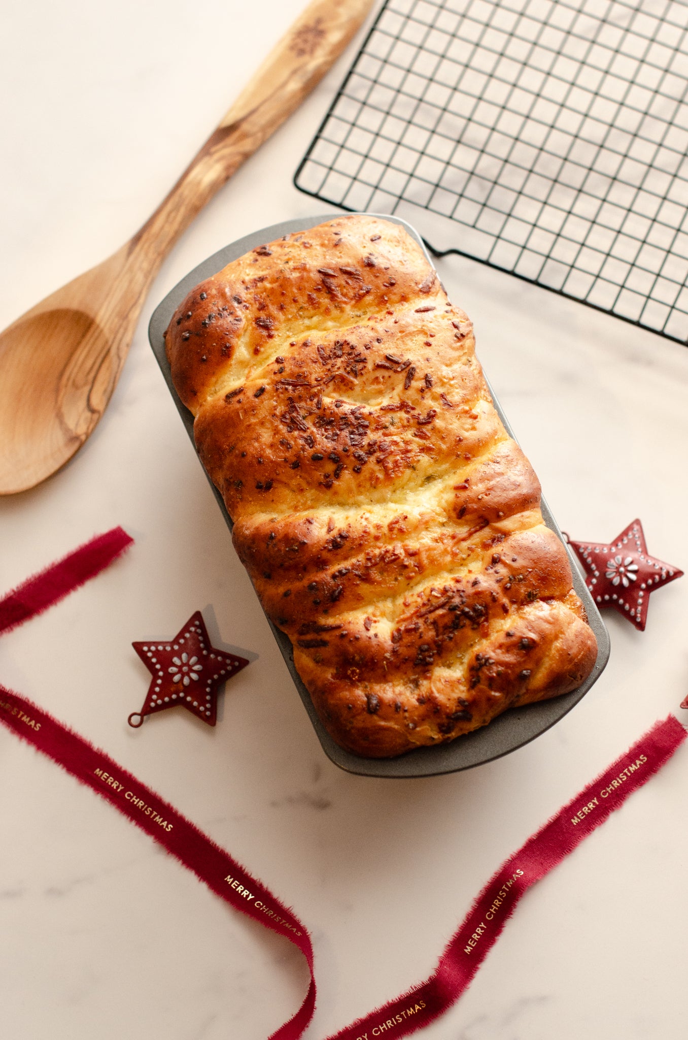 Golden garlic parmesan butter bread loaf topped with herbs in a pan on a marble surface with Christmas decorations, a metal cooling rack, and a wooden spoon, baked fresh by Chillable Bake Shop in Darien, Wisconsin.