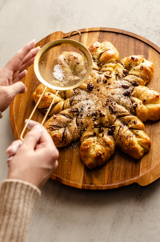 Chocolate pecan snowflake bread on a wooden board with a hand using a sieve to sprinkle powdered sugar on top, baked fresh for the holidays by Chillable Bake Shop in Darien, WI.