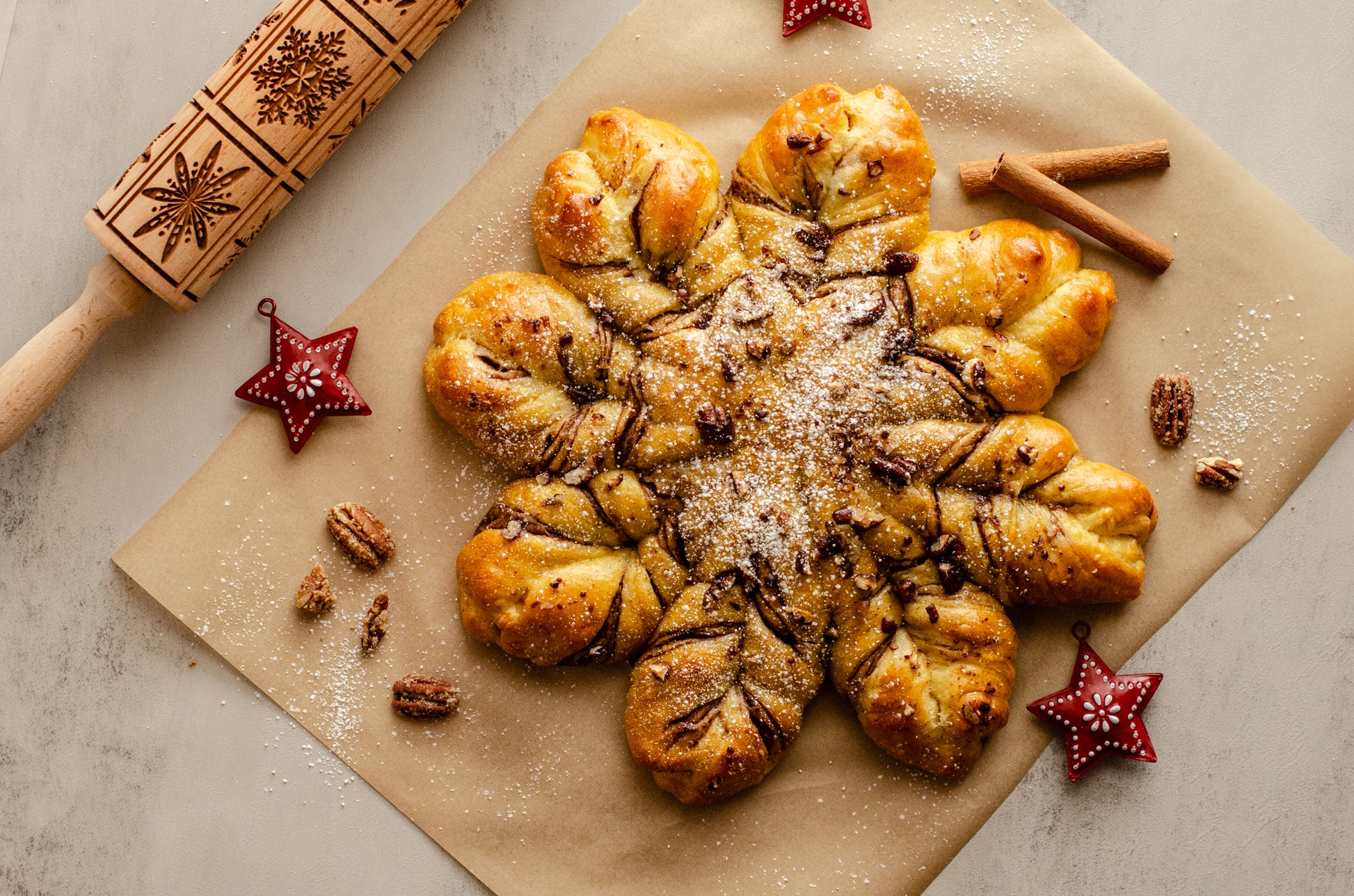 Decorative pastry shaped like a snowflake with layers of hazelnut chocolate and a dusting of powdered sugar on brown paper with cinnamon sticks and star-shaped decorations, baked fresh for the holidays by Chillable Bake Shop in Darien, WI.