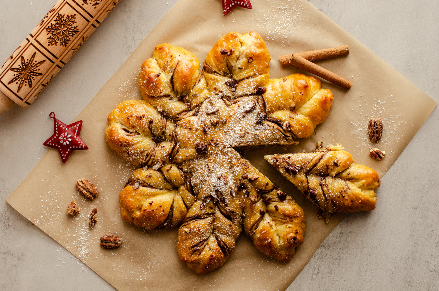 Decorative pastry shaped like a snowflake with layers of hazelnut chocolate and a dusting of powdered sugar - showing a slice cut out on brown paper with cinnamon sticks and star-shaped decorations, baked fresh for the holidays by Chillable Bake Shop in Darien, WI.
