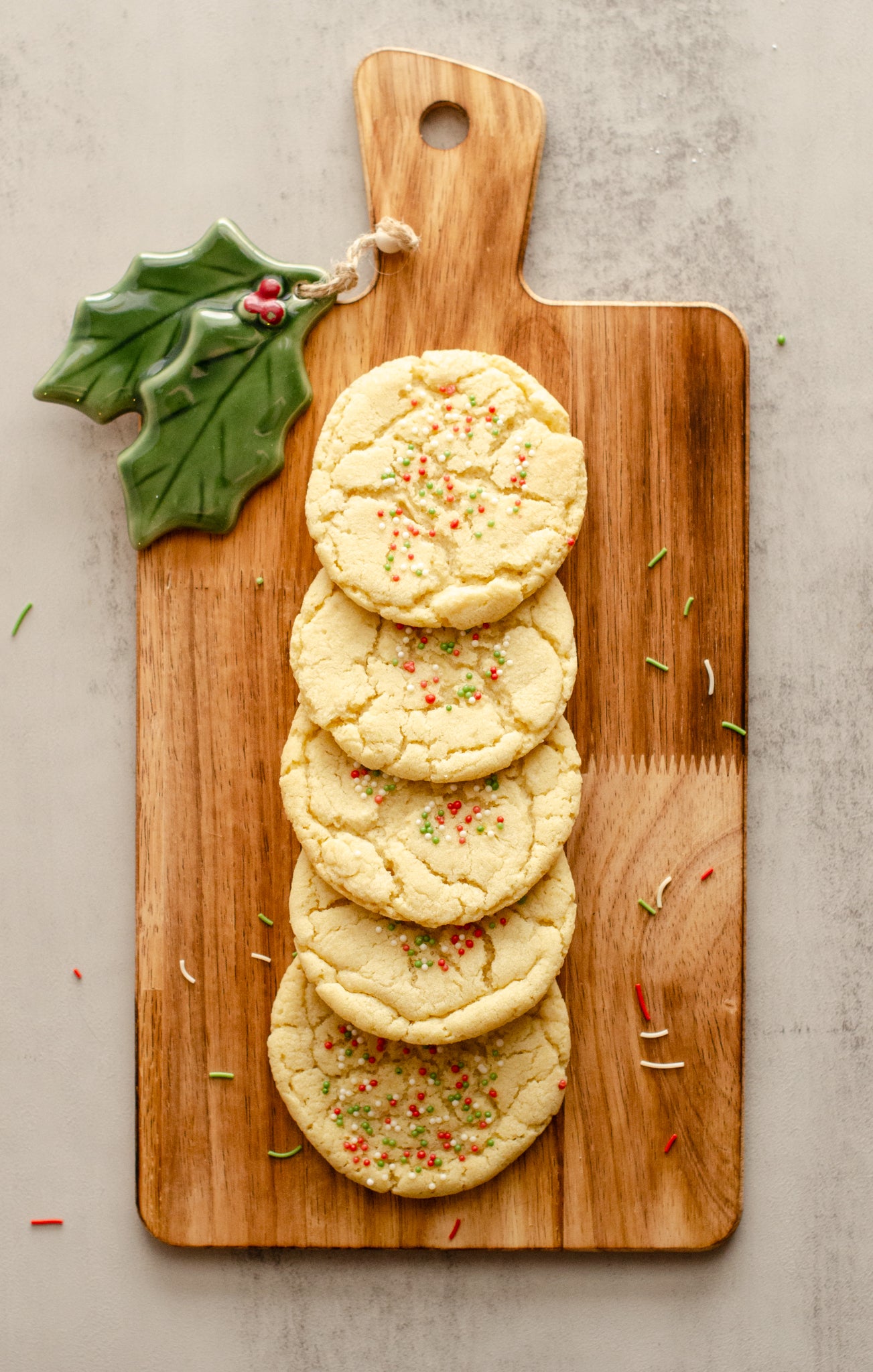 Holiday-themed sugar cookies with festive sprinkles on a wooden cutting board with a holly leaf ornament in the upper left corner, baked fresh by Chillable Bake Shop in Darien, Wisconsin.