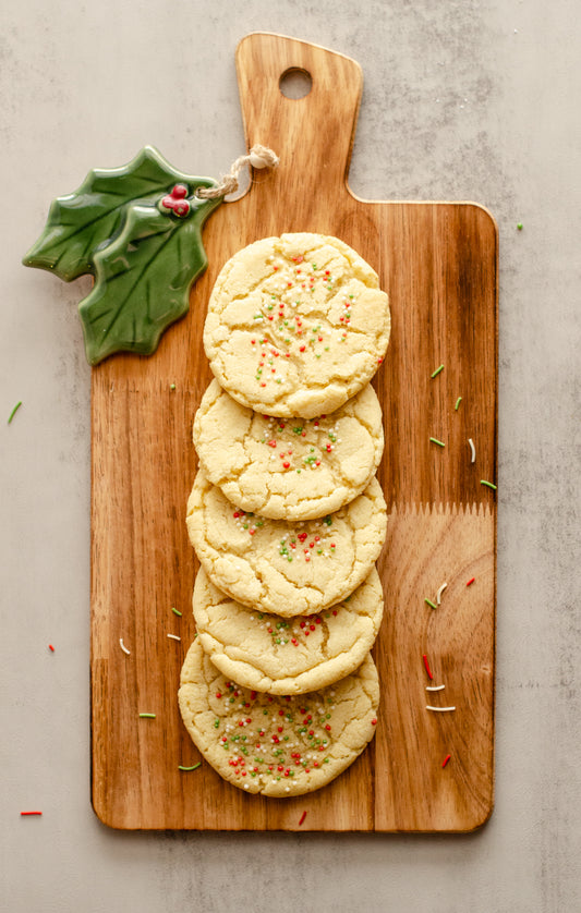 Holiday-themed sugar cookies with festive sprinkles on a wooden cutting board with a holly leaf ornament in the upper left corner, baked fresh by Chillable Bake Shop in Darien, Wisconsin.