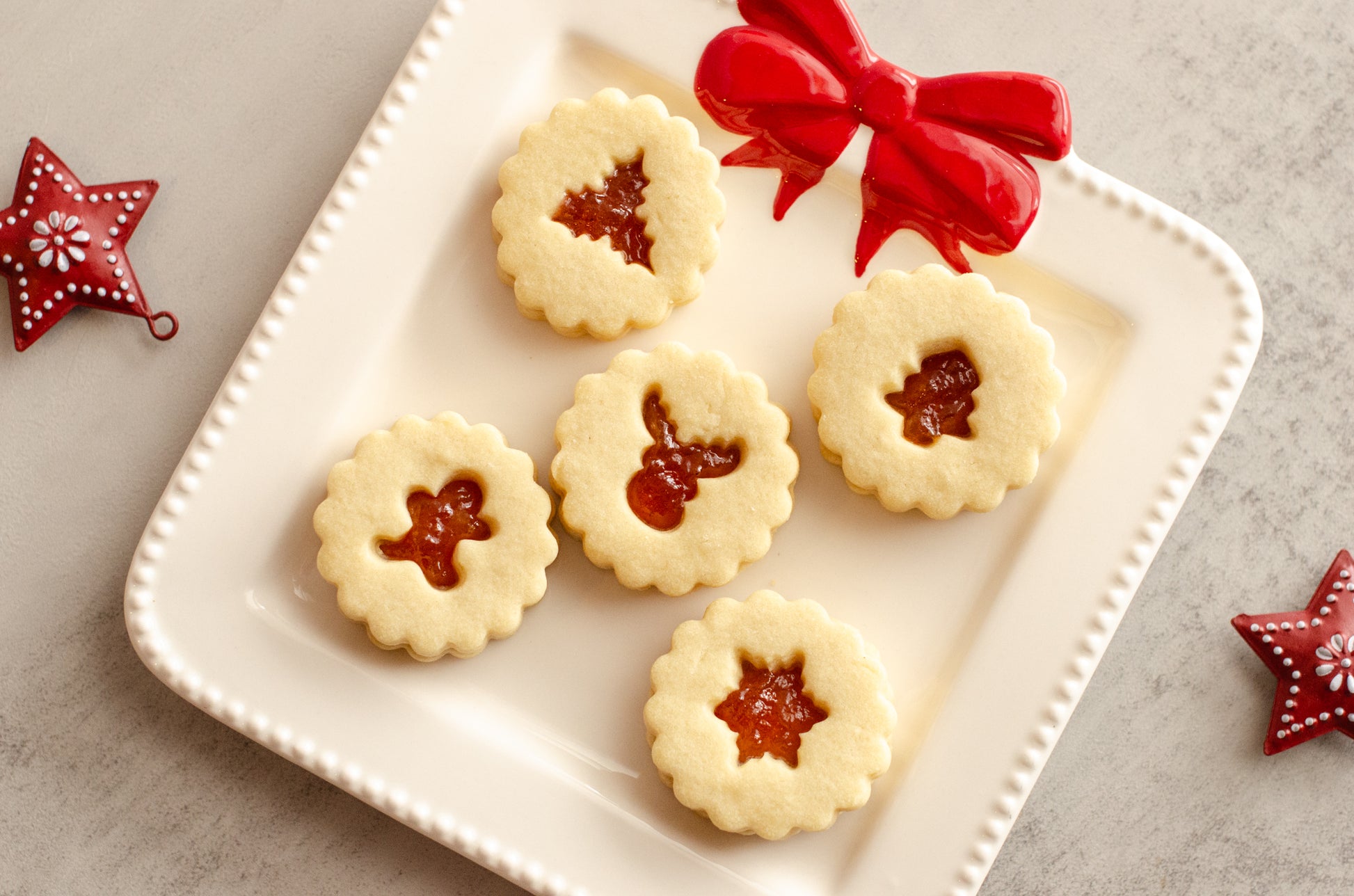 Decorative plate with round Peach Nutmeg Shortbread Linzer cookies featuring jam centers and holiday cut-outs, surrounded by festive stars on a neutral background. Baked fresh by Chillable Bake Shop in Darien, WI. Available for pickup and deliver.