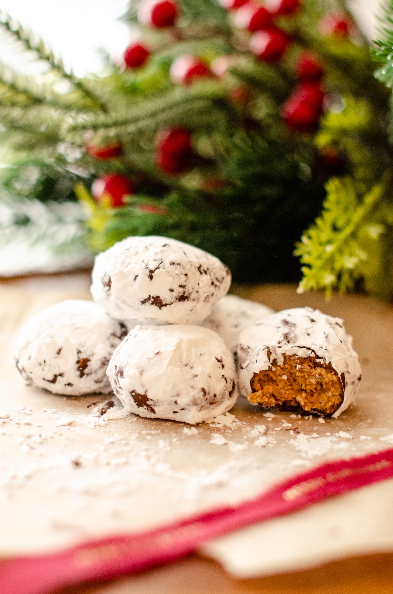 Stacked Puppy Chow Snowball Truffles rolled in powdered sugar, featuring one showing the crunchy peanut butter center with Christmas greenery and red berries in the background, handmade by Chillable Bake Shop in Darien, WI.