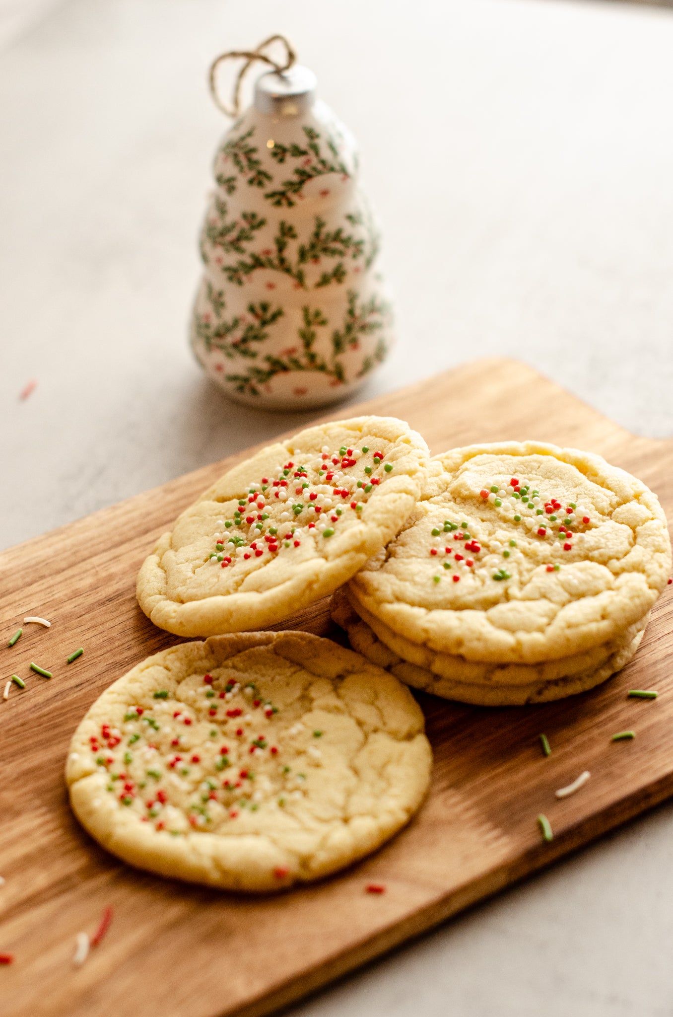 Three holiday themed sugar cookies with festive sprinkles on a wooden board with a decorative Christmas tree ornament in the background, baked fresh by Chillable Bake Shop in Darien, Wisconsin.