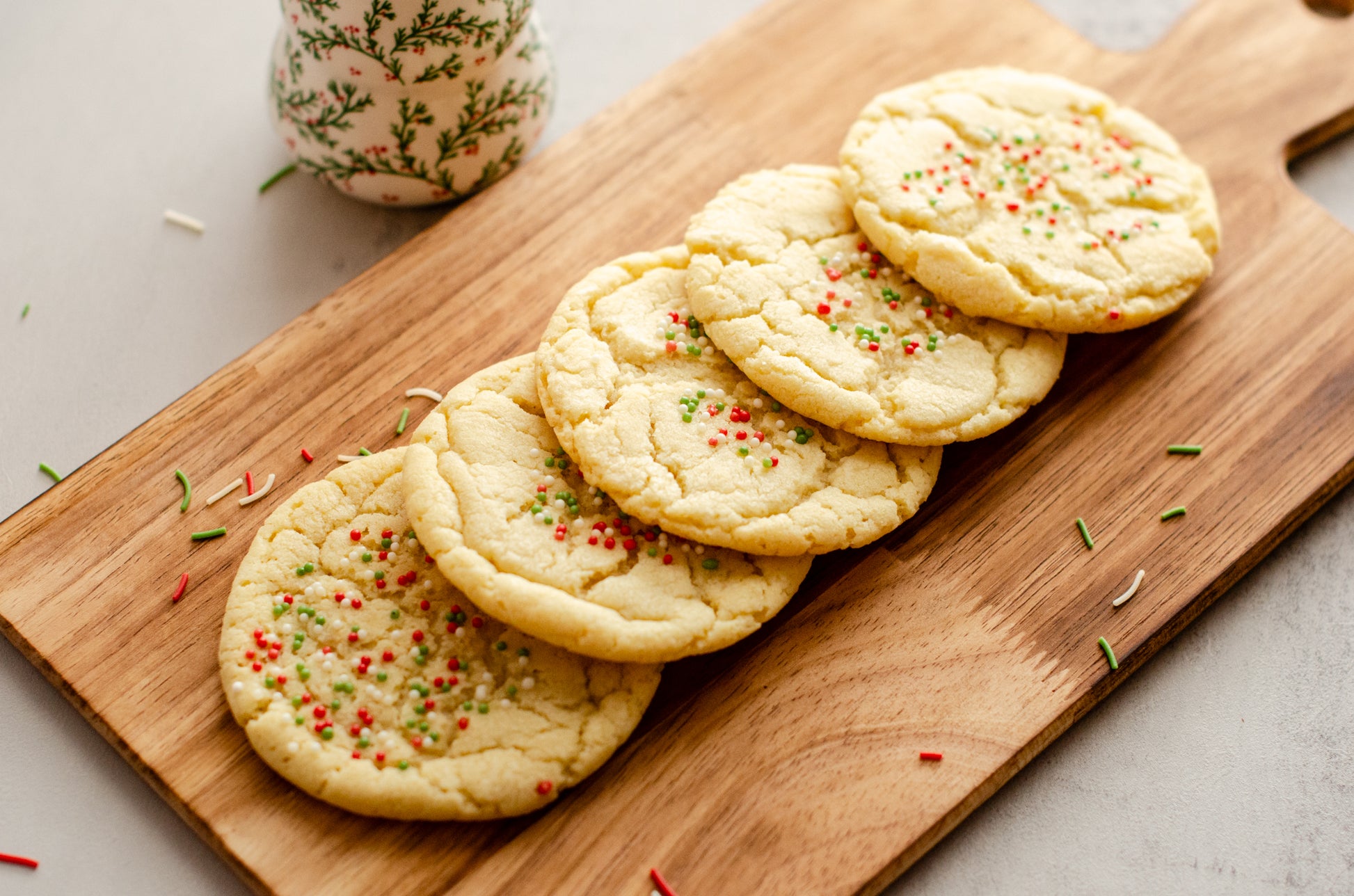 Three holiday themed sugar cookies with festive sprinkles on a wooden board with a decorative Christmas tree ornament in the background, baked fresh by Chillable Bake Shop in Darien, Wisconsin.