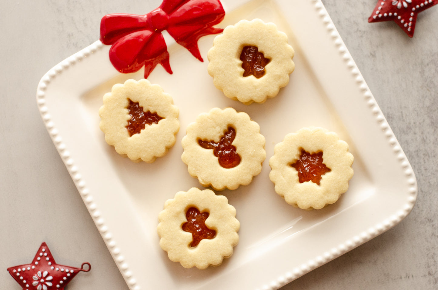 Peach nutmeg shortbread Linzer cookies with cutout centers on a white plate with a red bow, on a gray surface with Christmas decorations. Baked fresh by Chillable Bake Shop in Darien, WI. 