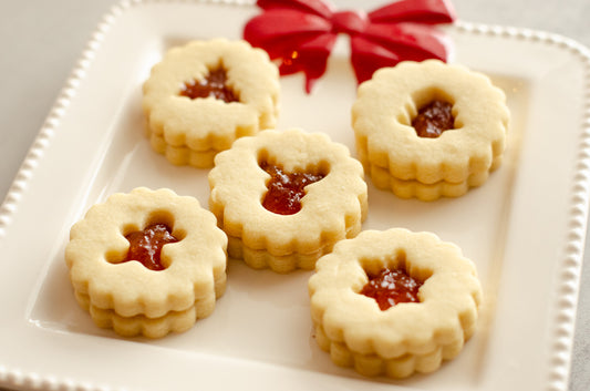 Peach nutmeg shortbread Linzer cookies with cutout centers on a white plate with a red bow, on a gray surface with Christmas decorations. Baked fresh by Chillable Bake Shop in Darien, WI. 