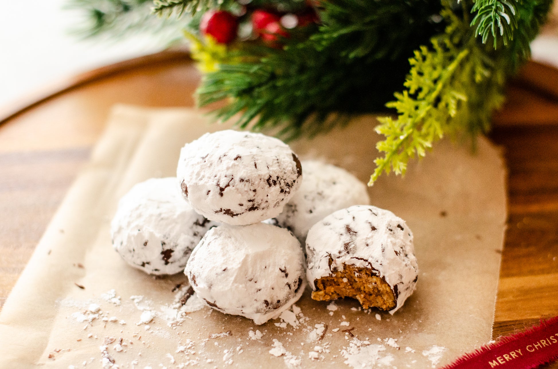 Stacked Puppy Chow Snowball Truffles rolled in powdered sugar, featuring one showing the crunchy peanut butter center with Christmas greenery and red berries in the background, handmade by Chillable Bake Shop in Darien, WI.