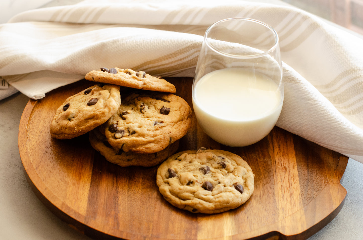 Stack of soft, chewy, chocolate chip cookies on a wooden surface with a glass of milk and a white cloth in the background , baked fresh by Chillable Bake Shop in Darien, Wisconsin.