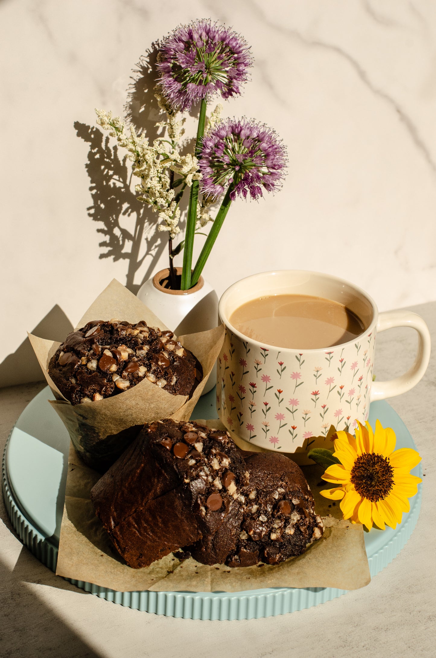 Two Mocha muffins with chocolate and espresso flavors in brown paper cups on a blue serving plate next to a cup of coffee in a flower patterned mug with a flower vase in the background, baked by Chillable Bake Shop in Darien, Wisconsin.