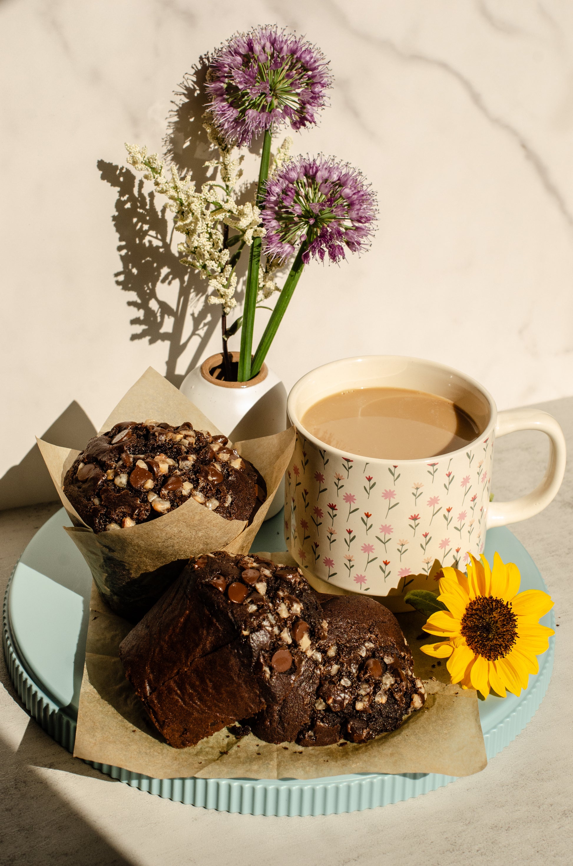 Two Mocha muffins with chocolate and espresso flavors in brown paper cups on a blue serving plate next to a cup of coffee in a flower patterned mug with a flower vase in the background, baked by Chillable Bake Shop in Darien, Wisconsin.