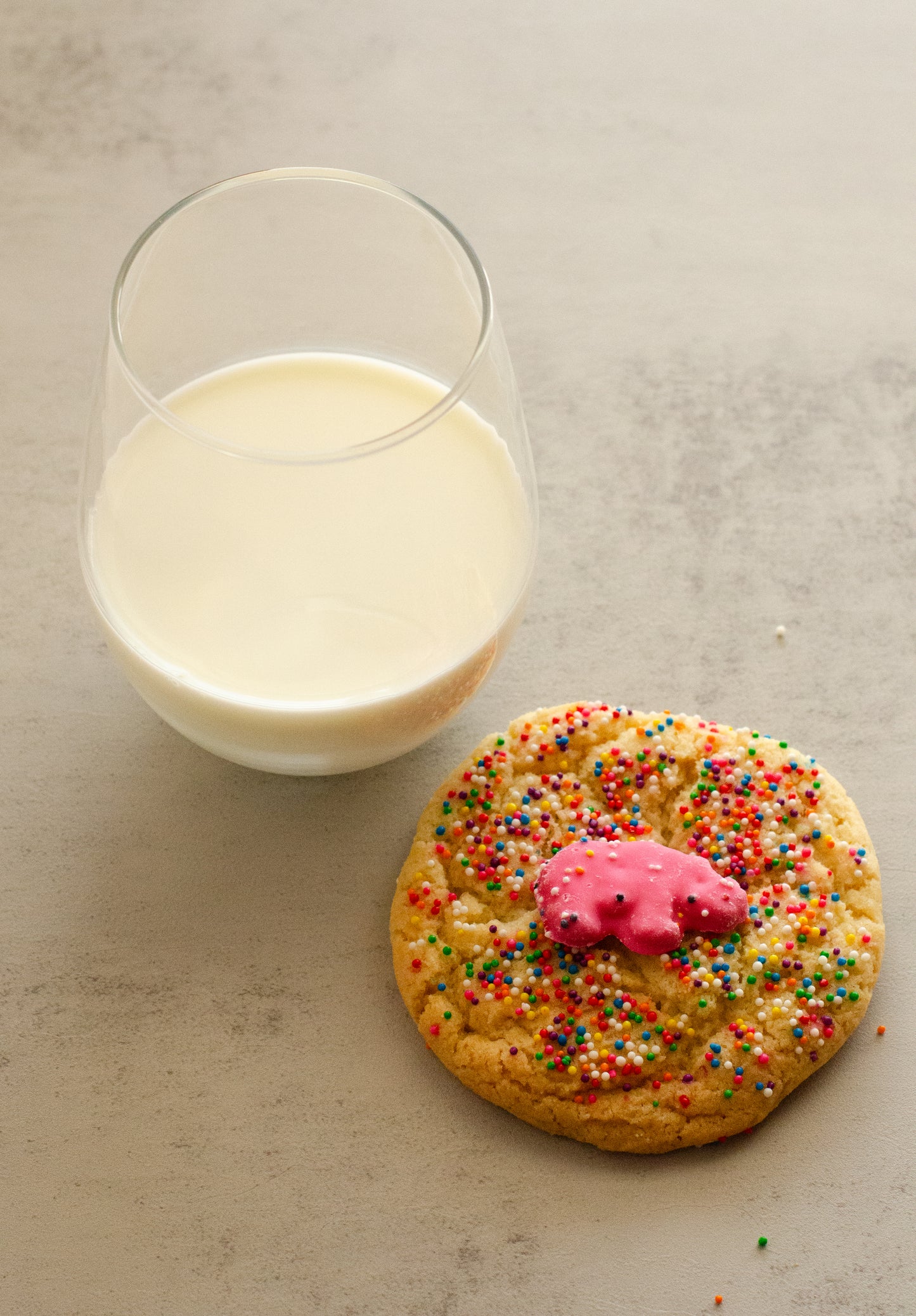 Glass of milk next to a sugar cookie topped with a pink frosted animal cookie and sprinkles on a neutral background, baked fresh by Chillable Bake Shop in Darien, Wisconsin.