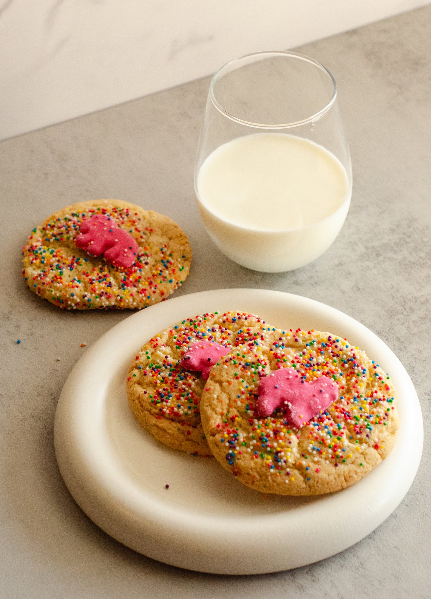Three sugar cookies, each topped with a pink frosted animal cookie and colorful sprinkles on a white plate, accompanied by a glass of milk, baked fresh by Chillable Bake Shop in Darien, Wisconsin.