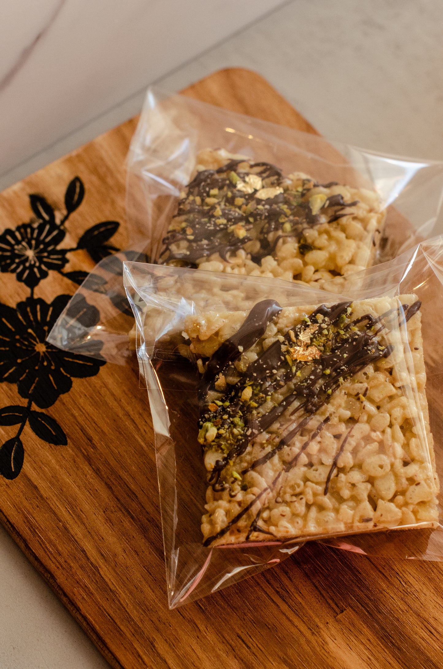 Two packages of Pistachio Chocolate Krispie treats with chopped pistachios on a wooden cutting board with a floral design, handmade in Darien, Wisconsin.